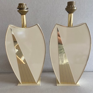 May include: Two matching cream-colored table lamps with a shield-like design. Each lamp features a gold-colored metal base, trim, and a diagonal accent. The lamps have a brass-colored neck and a bulb socket at the top.