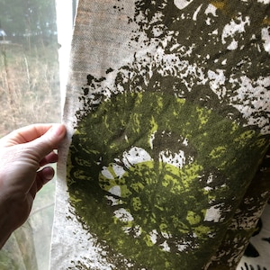 Puede incluir: Un primer plano de una cortina de tela con fondo blanco y un patrón abstracto verde y marrón. La tela tiene una apariencia texturizada, similar al lino.