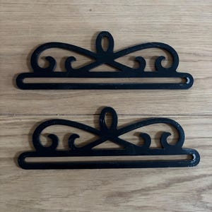 Könnte beinhalten: Zwei schwarze Metall-Regalhalterungen mit einem dekorativen Schnörkeldesign. Die Halterungen sind für die Wandmontage konzipiert und haben eine flache Oberfläche, auf der ein Regal ruhen kann.