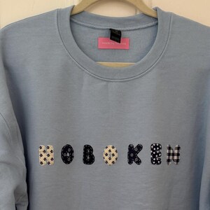May include: A light blue crewneck sweatshirt featuring the word HOBOKEN appliqued across the chest. Each letter is crafted from fabric with varying patterns, including black and white polka dots, gingham checks, and floral prints.