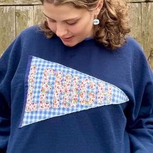 Könnte beinhalten: Ein marineblauer Pullover mit einem blauen und weißen karierten Wimpel, der auf der Vorderseite aufgenäht ist. Der Wimpel trägt das Wort "LEHIGH", das in Weiß mit floralen Akzenten gestickt ist.