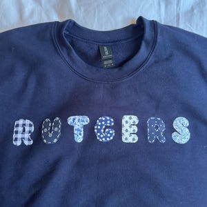 Może przedstawiać: Granatowa bluza z napisem "RUTGERS" na piersi. Litery wykonane są z różnych wzorzystych tkanin, w tym kratki, kwiatów i kropek. Bluza wykonana jest z miękkiego materiału.