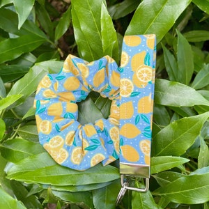 Op de afbeelding: Een set van een scrunchie en een polsbandje met citroenmotief. De items hebben een lichtblauwe achtergrond met gele citroenen en groene bladeren. De sleutelhanger heeft een zilverkleurige metalen sluiting.