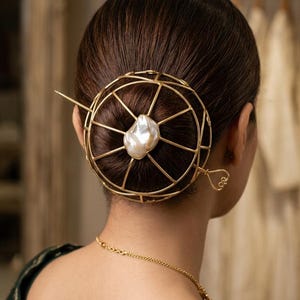 May include: A gold-colored hair accessory featuring a spherical cage design with a large, irregular pearl at the center. The hair piece is secured with a long pin and has a decorative loop detail. The accessory is worn in a bun.
