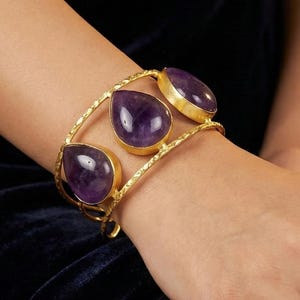 Peut inclure: Bracelet manchette doré orné de trois pierres précieuses violettes en forme de goutte d'eau. Le bracelet présente un motif texturé et est porté au poignet, mettant en valeur le style élégant du bijou. Les pierres sont serties dans des chatons dorés.