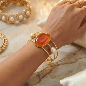 May include: Gold-coloured bracelet with a large, oval, orange gemstone and two white pearls. The open cuff design is accented with delicate gold lines. Other gold bracelets with pearls and gemstones are also visible.