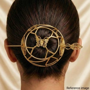 Puede incluir: Accesorio para el cabello dorado con un diseño circular con una mariposa en el centro. La pieza para el cabello tiene un alfiler largo y un extremo decorativo en forma de hoja, diseñado para asegurar el cabello.