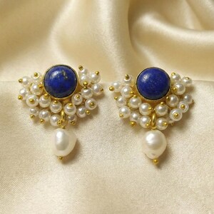 Peut inclure: Une paire de boucles d'oreilles dorées avec un centre en pierre précieuse bleue entouré de petites perles blanches. Chaque boucle d'oreille a une perle blanche pendante. Les boucles d'oreilles sont posées sur un fond crème.