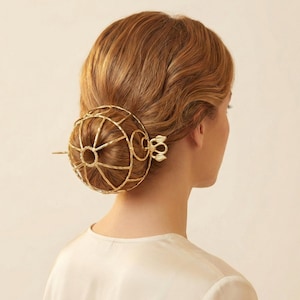 Könnte beinhalten: Ein goldfarbenes Haaraccessoire mit einem kugelförmigen Käfigdesign und einer dekorativen Nadel. Das Haarteil wird im Haar getragen, mit einem blütenartigen Akzent. Das Accessoire ist aus Metall.