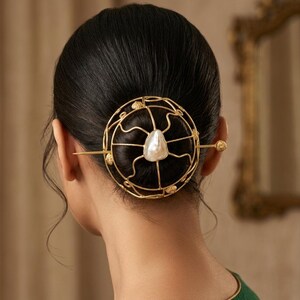 Könnte beinhalten: Ein goldfarbenes Haaraccessoire mit kreisförmigem Design, einer zentralen Perle und filigranen Drahtdetails. Das Haarteil wird mit einem langen Stift befestigt, was dem Haarstil einen Hauch von Eleganz verleiht. Das Accessoire wird im Nacken getragen.