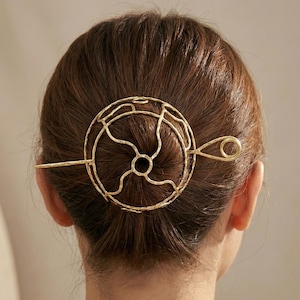 May include: A gold-colored hair pin with a circular design and a long, straight pin. The circular part has an open, abstract pattern. The hair accessory is worn in a bun, with the pin inserted through the hair.