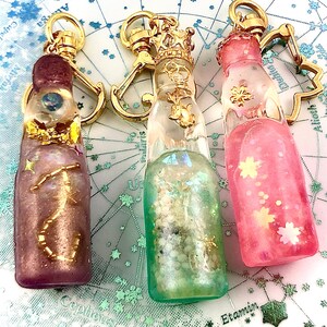 May include: Three glass bottle keychains with gold accents and different colored liquids inside. The bottles are filled with glitter and small decorative items. The keychains are attached to gold metal rings.