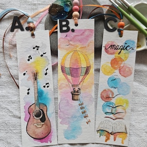 Op de afbeelding: Drie aquarel bladwijzers met decoratieve linten. Eén heeft een gitaar en muzieknoten, een andere een heteluchtballon, en de derde heeft het woord "magic" met kleurrijke ballonnen en een open boek. Elke bladwijzer is handgeschilderd.