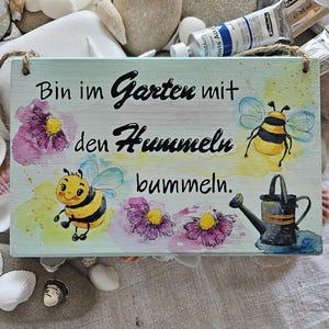 May include: A rectangular wooden sign with a light green background featuring hand-painted illustrations of two bumblebees, three purple flowers, and a black watering can. The text reads: Bin im Garten mit den Hummeln bummeln.