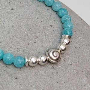 May include: A beaded bracelet featuring round, light blue gemstone beads alternating with polished silver-toned spherical beads and a single central silver-toned bead with a raised spiral design, arranged in a circular pattern on a gray surface.