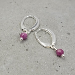 May include: A pair of silver leverback earrings with a small, faceted, magenta-colored gemstone dangling from each earring. The earrings are displayed on a gray surface.
