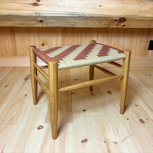 May include: A rectangular stool with four light brown wooden legs and a woven seat featuring a repeating chevron pattern in alternating strips of tan and reddish-brown material. The stool has horizontal support bars between the legs.
