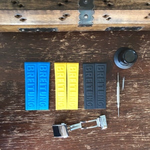 May include: Three pairs of watch straps, one blue, one yellow, and one black, with the word "BREITLING" printed on each strap. A silver watch buckle is also pictured.