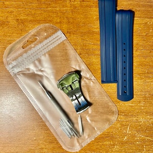 Puede incluir: Una bolsa de pl&aacute;stico transparente que contiene una correa de reloj plateada con una hebilla verde, una peque&ntilde;a herramienta plateada y varios pasadores plateados peque&ntilde;os. Dos correas de reloj azul oscuro est&aacute;n al lado de la bolsa.