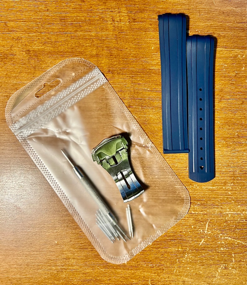 Puede incluir: Una bolsa de pl&aacute;stico transparente que contiene una correa de reloj plateada con una hebilla verde, una peque&ntilde;a herramienta plateada y varios pasadores plateados peque&ntilde;os. Dos correas de reloj azul oscuro est&aacute;n al lado de la bolsa.
