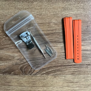 Puede incluir: Dos correas de reloj naranjas con una herramienta de correa de reloj plateada y pasadores de resorte en una bolsa de pl&aacute;stico transparente.