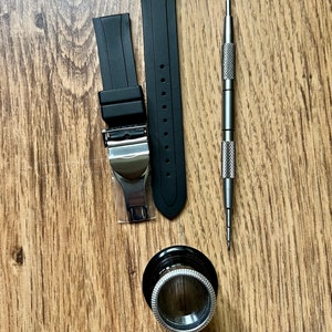 Puede incluir: Dos correas de reloj de goma negra con hebillas plateadas, una herramienta de correa de reloj plateada y una lupa plateada sobre una superficie de madera.