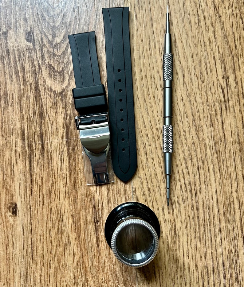 Puede incluir: Dos correas de reloj de goma negra con hebillas plateadas, una herramienta de correa de reloj plateada y una lupa plateada sobre una superficie de madera.