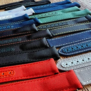 Puede incluir: Una colección de correas de reloj en varios colores, incluyendo negro, azul, verde y rojo. Las correas están hechas de un material tejido y presentan costuras en contraste y detalles de cuero. Para relojes.