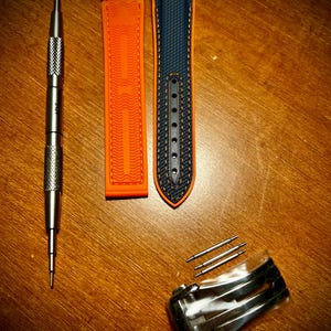 Puede incluir: Un surtido de accesorios para relojes, que incluyen una correa de reloj naranja y azul marino, una herramienta de reloj plateada, un cierre plateado y varios pasadores plateados. La correa del reloj est&aacute; hecha de un material texturizado.