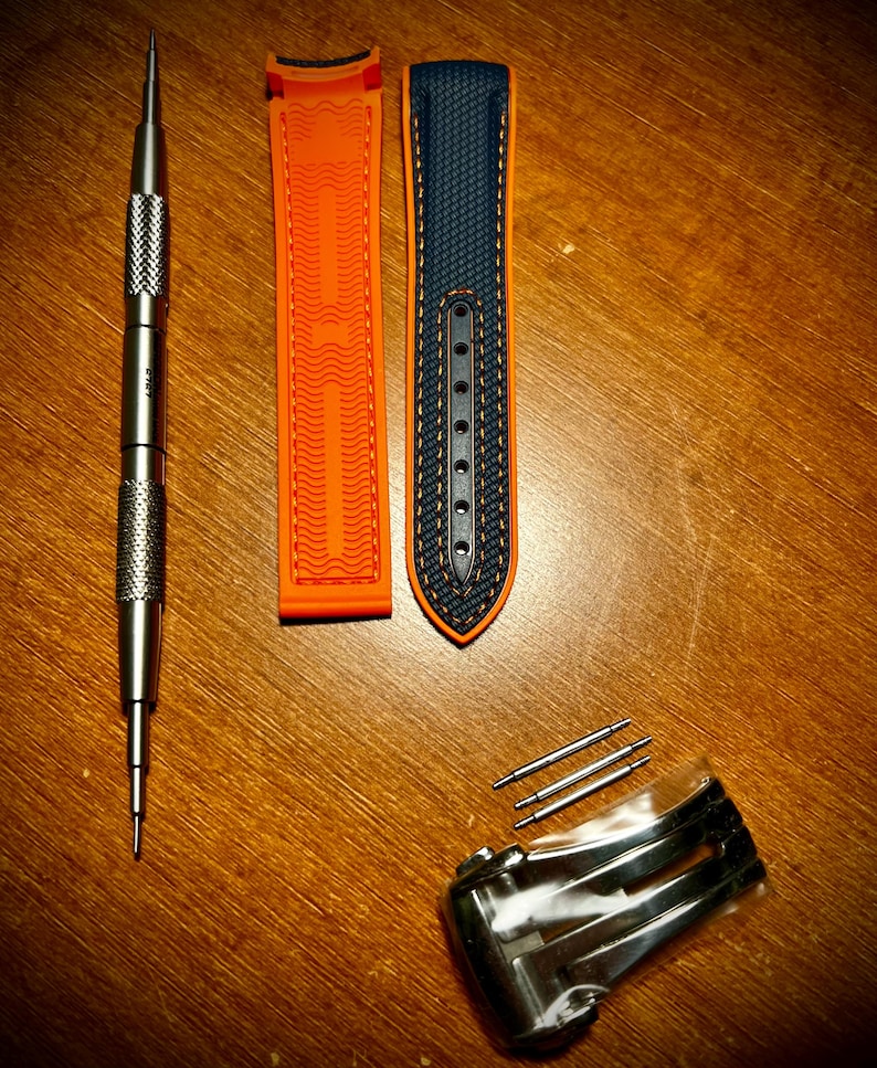 Puede incluir: Un surtido de accesorios para relojes, que incluyen una correa de reloj naranja y azul marino, una herramienta de reloj plateada, un cierre plateado y varios pasadores plateados. La correa del reloj est&aacute; hecha de un material texturizado.