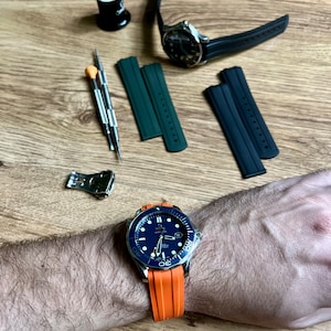 Puede incluir: Un reloj azul y plateado con una correa de caucho naranja. El reloj tiene una esfera negra con marcas blancas y una ventana de fecha en la posici&oacute;n de las 3 en punto. El reloj est&aacute; en la mu&ntilde;eca de una persona.