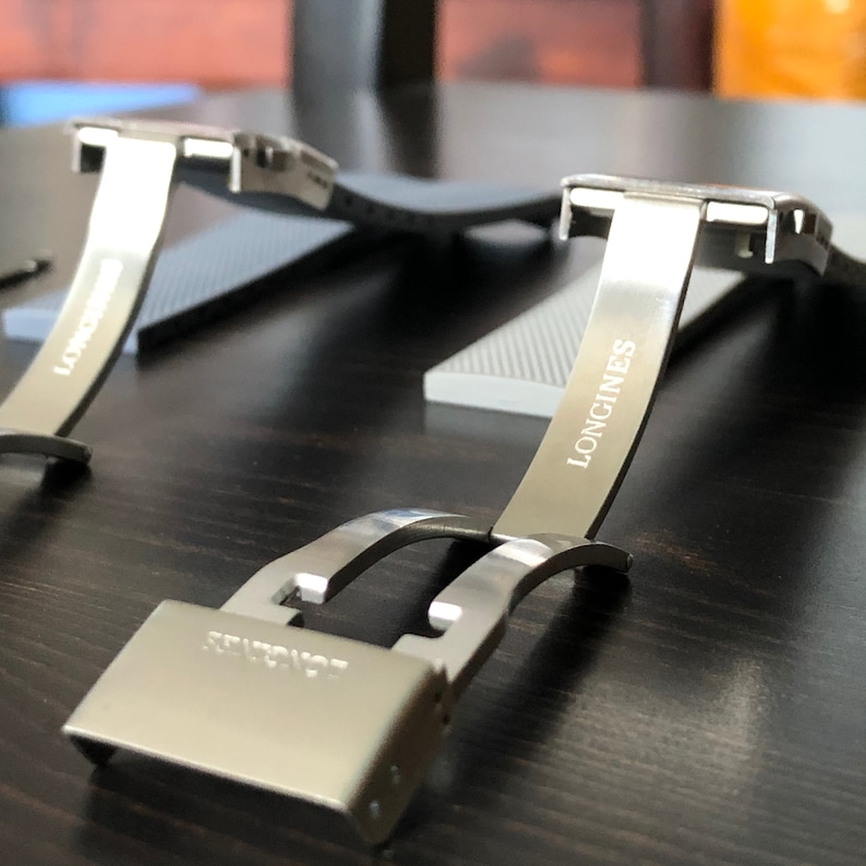 Puede incluir: Una hebilla de correa de reloj plateada con la palabra "LONGINES" grabada en el lateral. La hebilla es de metal y tiene un acabado pulido.