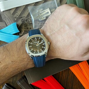May include: A silver wristwatch with a dark brown face and a blue rubber strap is shown on a wrist. The watch has white numerals and hands. Various watch straps in white, blue, green, red, and orange are visible.