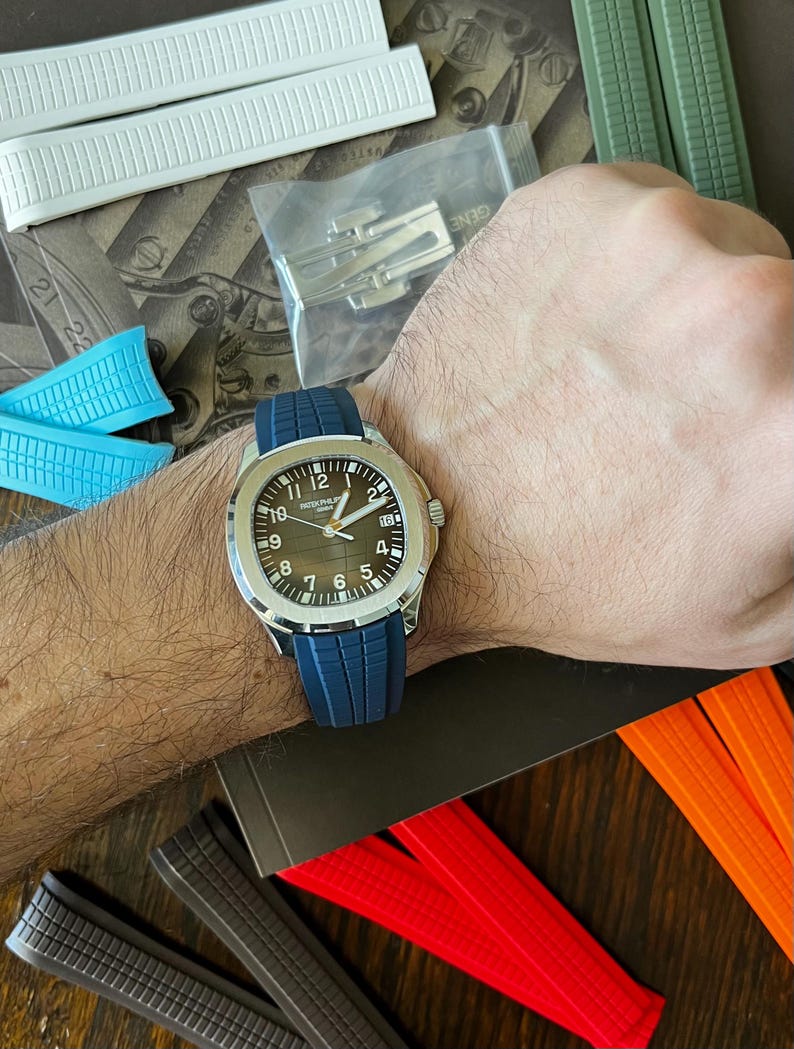 May include: A silver wristwatch with a dark brown face and a blue rubber strap is shown on a wrist. The watch has white numerals and hands. Various watch straps in white, blue, green, red, and orange are visible.
