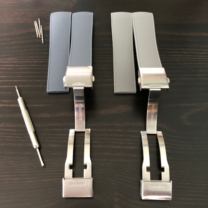 Puede incluir: Dos correas de reloj de silicona grises con hebillas de metal plateado. Las hebillas tienen la palabra "LONGINES" grabada.