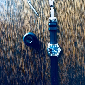 Puede incluir: Un reloj negro con una banda plateada y una correa de caucho negra. El reloj tiene una esfera negra con n&uacute;meros y manecillas blancas. El reloj est&aacute; sobre una superficie de madera con una peque&ntilde;a herramienta y una hebilla de correa de reloj.