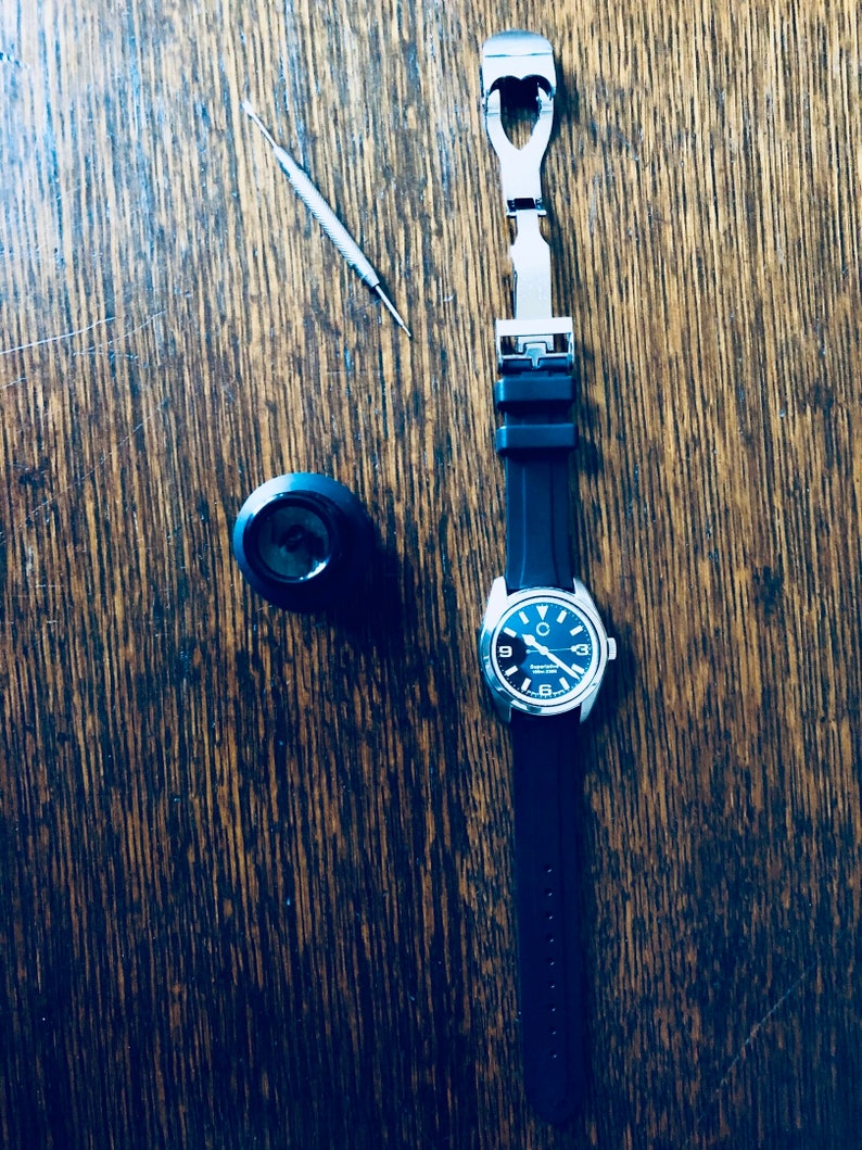 Puede incluir: Un reloj negro con una banda plateada y una correa de caucho negra. El reloj tiene una esfera negra con n&uacute;meros y manecillas blancas. El reloj est&aacute; sobre una superficie de madera con una peque&ntilde;a herramienta y una hebilla de correa de reloj.