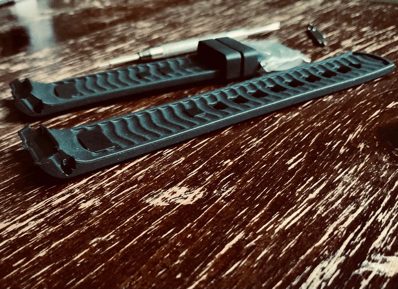 May include: Two black watch bands with a textured surface. The bands are laid out on a wooden surface.