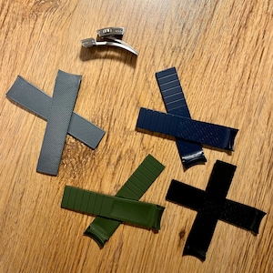 Puede incluir: Cuatro correas de reloj de diferentes colores y materiales. Una correa gris texturizada, una correa verde oscuro, una correa negra y una correa azul oscuro. Las correas est&aacute;n dispuestas en c&iacute;rculo sobre una superficie de madera.