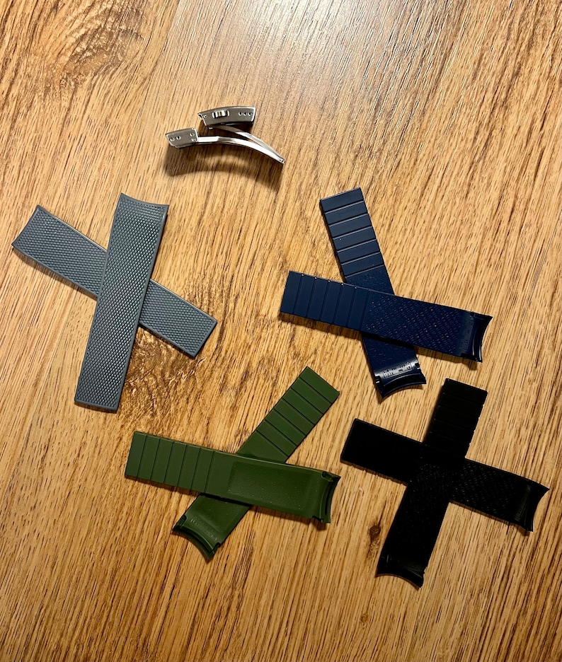 Puede incluir: Cuatro correas de reloj de diferentes colores y materiales. Una correa gris texturizada, una correa verde oscuro, una correa negra y una correa azul oscuro. Las correas est&aacute;n dispuestas en c&iacute;rculo sobre una superficie de madera.