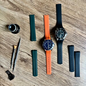 Puede incluir: Dos relojes de pulsera con esferas negras y agujas plateadas en correas de caucho de diferentes colores. Las correas son naranja y verde. Tambi&eacute;n hay cuatro correas de reloj adicionales en negro y verde.