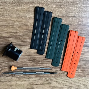 Puede incluir: Tres correas de reloj en negro, verde y naranja. Las correas son de goma y tienen una superficie texturizada. Las correas est&aacute;n dispuestas sobre una superficie de madera.