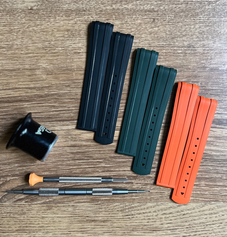 Puede incluir: Tres correas de reloj en negro, verde y naranja. Las correas son de goma y tienen una superficie texturizada. Las correas est&aacute;n dispuestas sobre una superficie de madera.