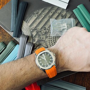 May include: A silver watch with a green face and an orange strap is displayed, surrounded by various watch straps in colours such as grey, green, and white. The watch is on a wrist.