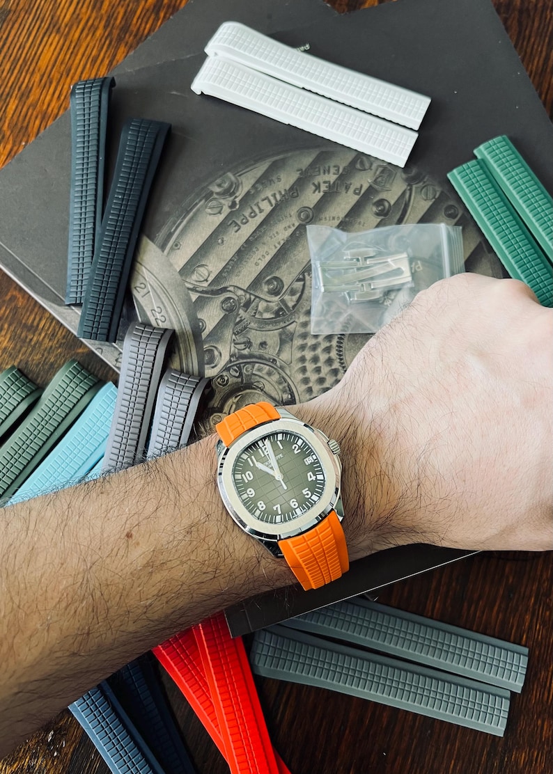May include: A silver watch with a green face and an orange strap is displayed, surrounded by various watch straps in colours such as grey, green, and white. The watch is on a wrist.