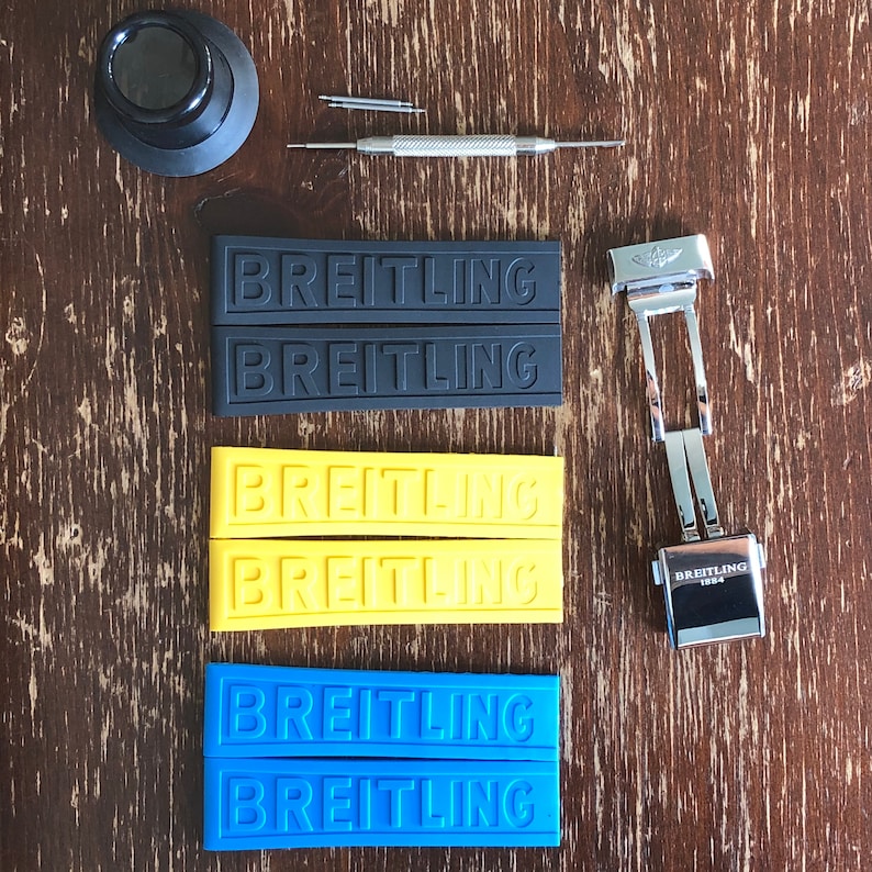 May include: A set of four watch straps with the word "Breitling" embossed on each strap. The straps are black, yellow, and blue. A silver watch band clasp is also pictured.