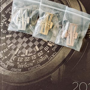 May include: Three watch clasps in clear plastic bags. The clasps are in silver, gold, and rose gold tones. The background features a watch face design with city names and the text "2021 COLLECTION PATEK PHILIPPE."