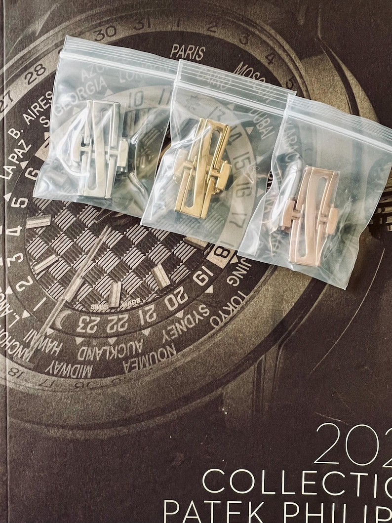 May include: Three watch clasps in clear plastic bags. The clasps are in silver, gold, and rose gold tones. The background features a watch face design with city names and the text "2021 COLLECTION PATEK PHILIPPE."