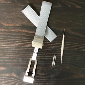 Puede incluir: Un cierre de pulsera de reloj plateado con dos correas de reloj grises y una herramienta para quitar los pasadores del reloj.