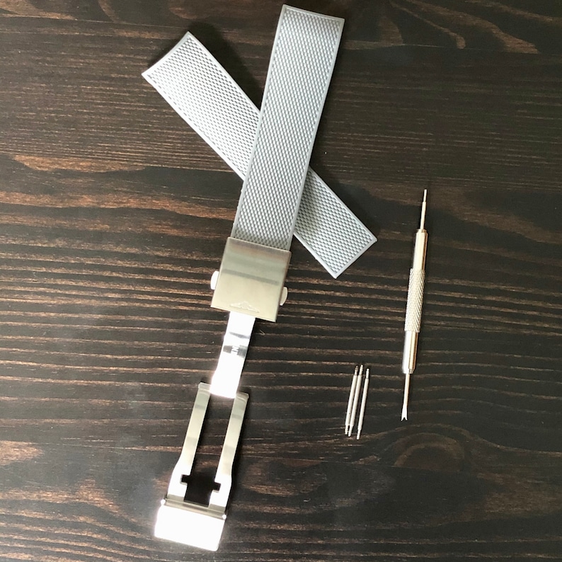 Puede incluir: Un cierre de pulsera de reloj plateado con dos correas de reloj grises y una herramienta para quitar los pasadores del reloj.